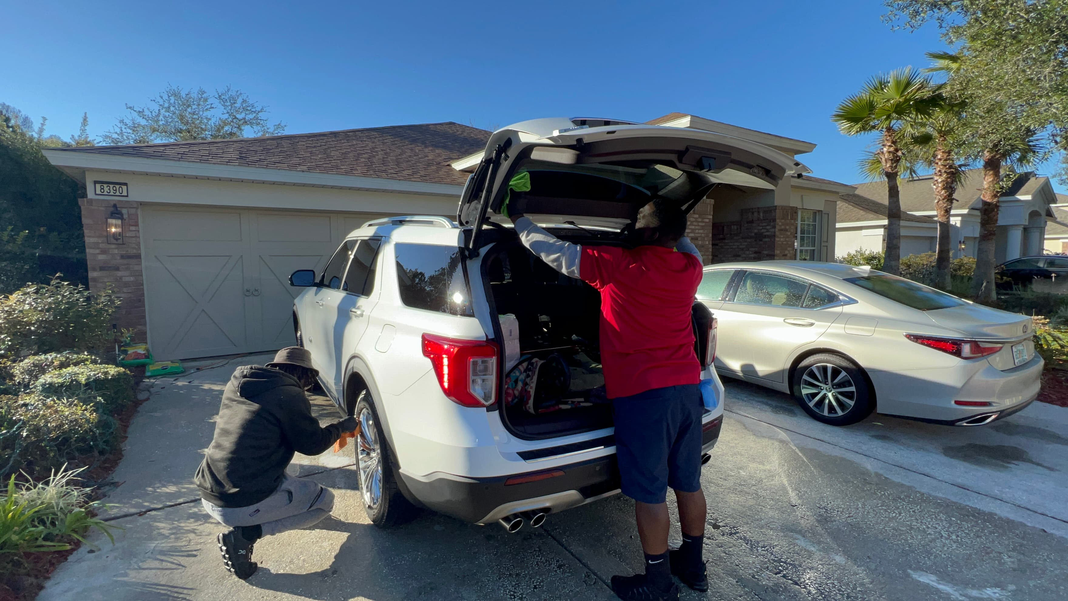 Two men cleaning a white SUV in a sunny driveway, wiping the trunk and wheel.