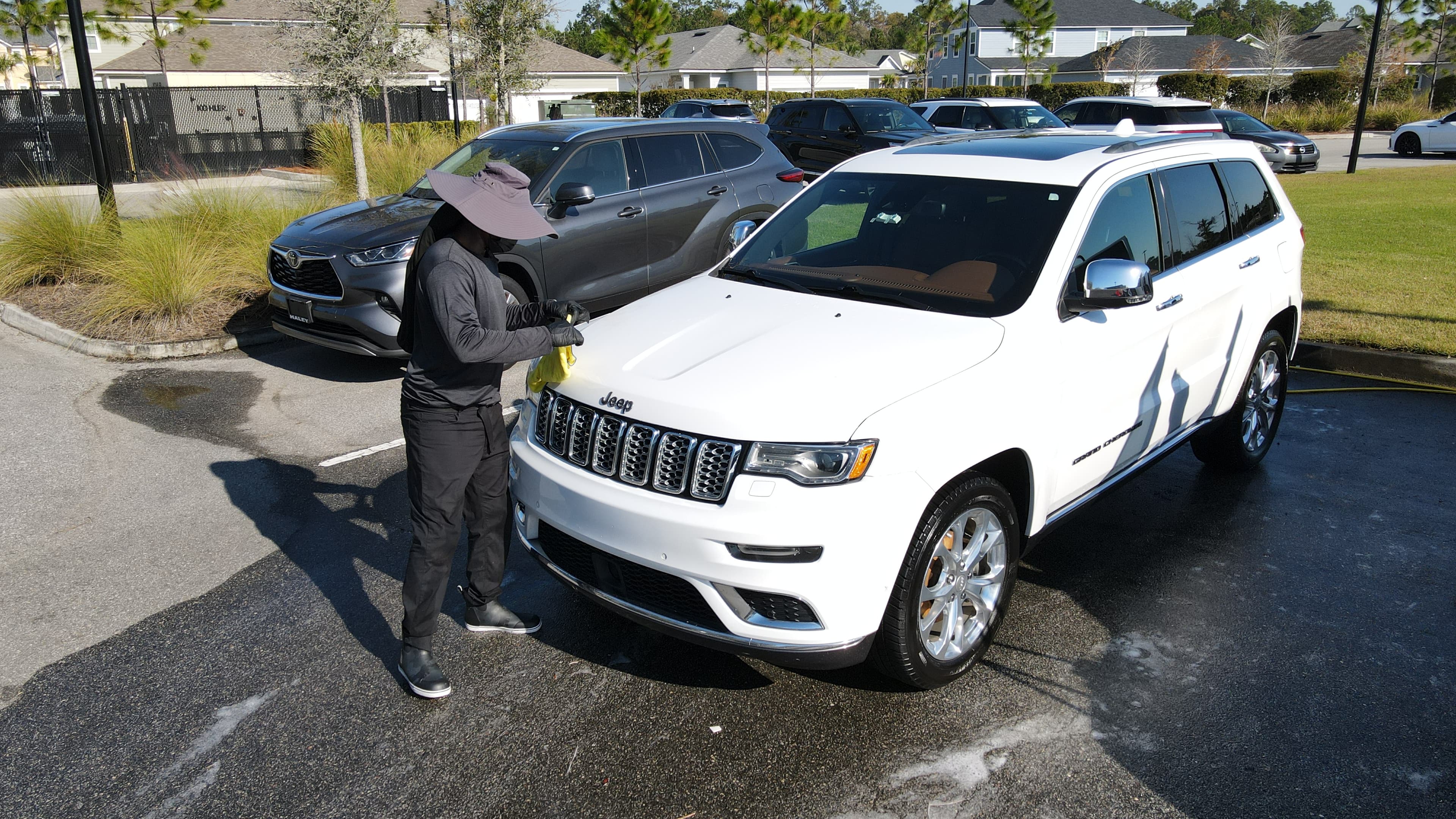Person in a sun hat wipes a white Jeep's hood with a yellow microfiber cloth.